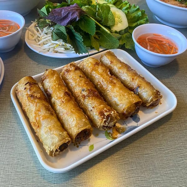 Five crispy Vietnamese imperial rolls on a white plate with fresh herbs and dipping sauce.
