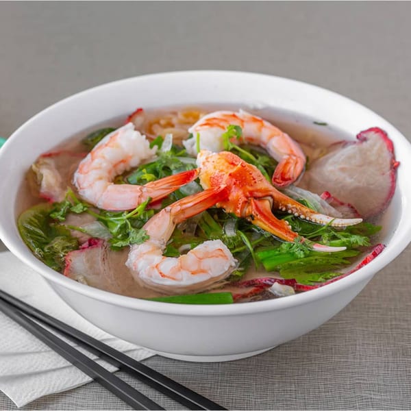 A bowl of Vietnamese pho with shrimp, crab, and pork, garnished with fresh herbs.